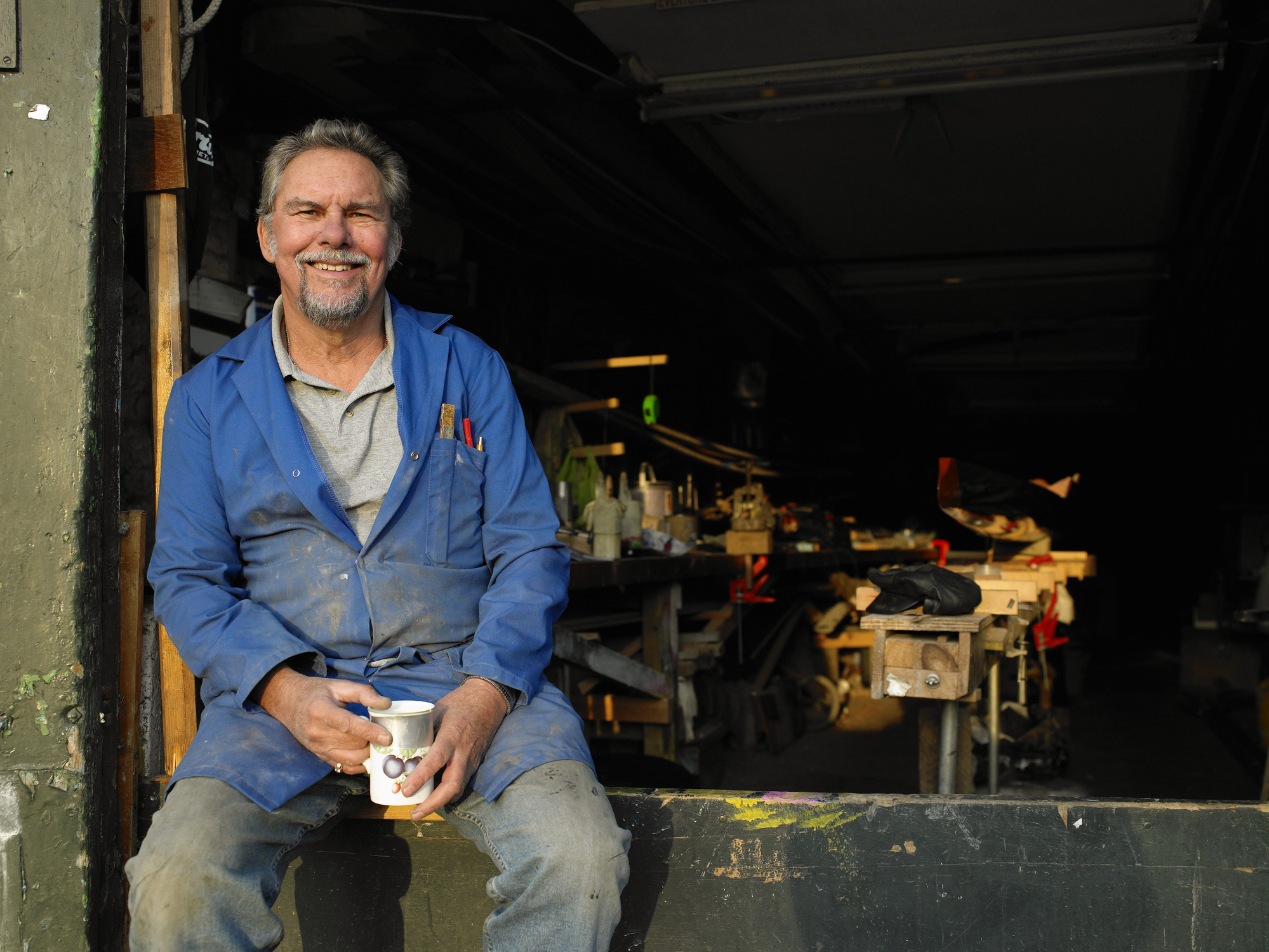 Senior male boat builder in workshop holding mug, smiling, portriat