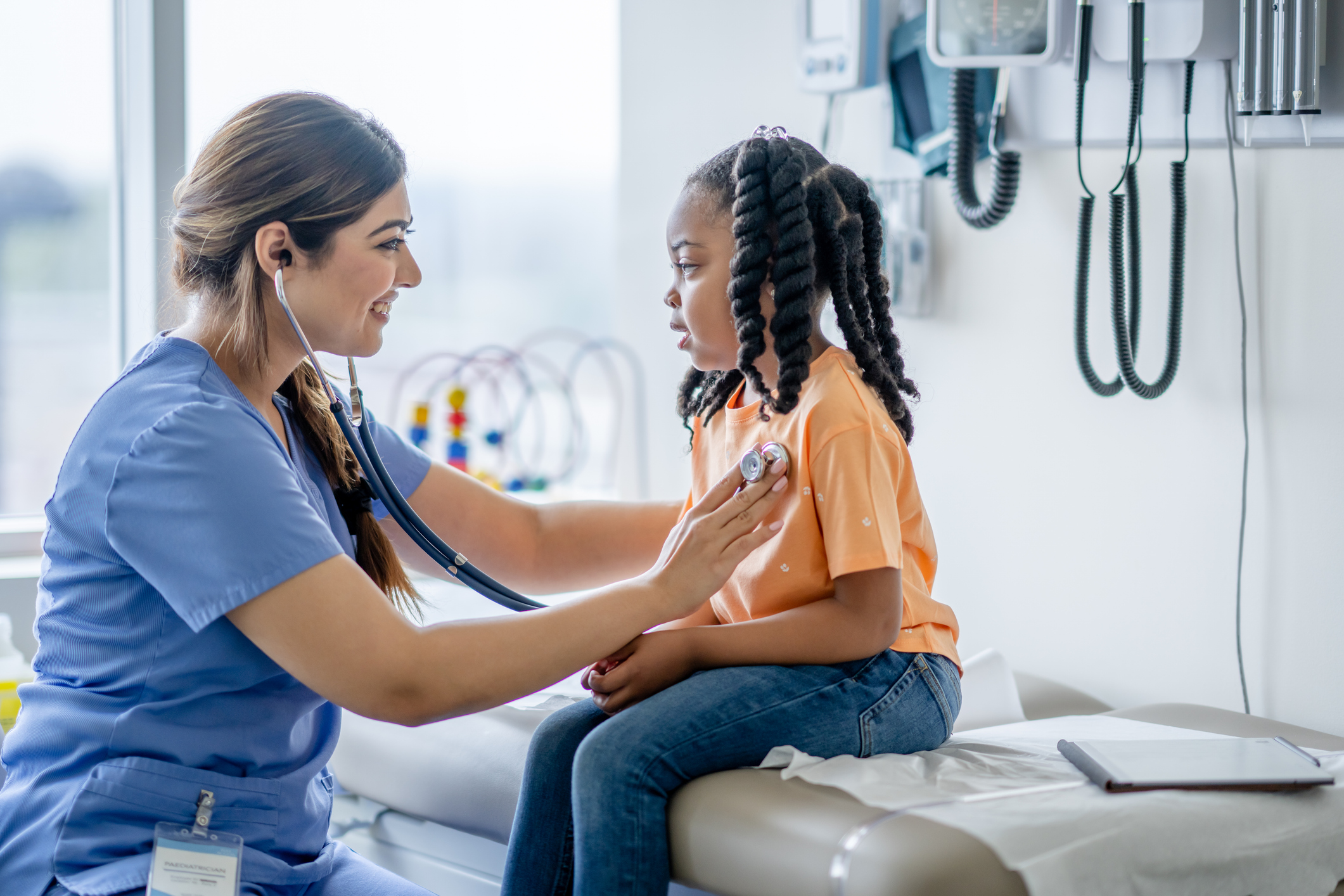 A sweet little girl of African decent, sits up on an exam table during a routine check-up. She is dressed casually and is smiling gently as her doctor leans in with her stethoscope to listen to her heart.
